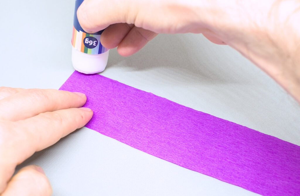 untando pegamento en barra en un lado de tira de papel crespon purpura
